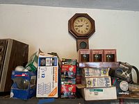 Photo of stacked boxes of light bulbs on a shelf below a wall clock, showing various brands and types including GE, Sylvania, and halogen bulbs.