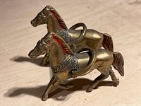 Close-up photo of two brass-colored metal horses with red painted manes, tails, harnesses, and rings on their backs on a wooden surface.