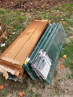 Stack of green window shutters with packaging, placed outside on grass with autumn leaves around.