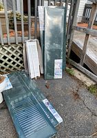 Photo showing multiple vinyl shutters stacked against a wooden deck, both green and white shutters visible