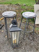 Two weathered metal stools with round seats and a hanging black metal outdoor light fixture with glass panels placed on gravel ground.