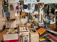 Wide angle photo showing various tools, clamps, boxes with light bulbs, sandpaper, saw blades, and other tool accessories on a pegboard and table.