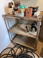 Photo of stainless steel utility cart with wheels, some hardware boxes and items on top, two caster wheels on middle shelf, and coiled cables on floor nearby.