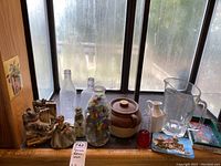 Overview photo showing entire lot on window sill including figurines, bottles, ceramic pot, glass pitcher and ashtray