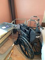 Black foldable wheelchair, rolling bed table, walker with tray, and toilet seat in a room with peach walls and wooden floor.