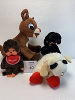Photo showing all four plush animals: Monchhichi doll, Rudolph reindeer, black dog, and Lambchop sheep, placed on a white surface with lot marker.