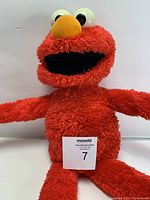 Front view of the red plush Elmo doll sitting upright with arms extended and a tag labeled '7' at the base.