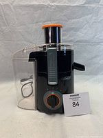 Front view of black Omega juicer with orange accents, visible transparent pulp container and power cord.