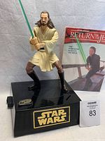 Front view of battery-operated Qui Gon Jinn figure standing on black base with Star Wars logo, holding green lightsaber. Return of the Jedi storybook placed behind figure.
