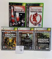 Five Xbox game cases displayed in two rows. Top row includes Rainbow Six Lockdown and Rainbow Six Critical Hour. Bottom row includes Ghost Recon 2, Splinter Cell (Platinum Hits), and Ghost Recon (Platinum Hits). Games are for Xbox and published by Ubisoft.