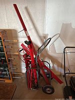 View of unassembled ceiling finishing equipment showing red metal parts stacked and chained on a wheeled cart with handle, stored indoors against a wall next to a foosball table.