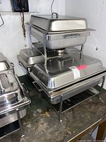 Photo showing stacked chafing trays with lids, frames, and Sterno holders underneath on a metal countertop.