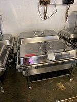 Three stainless steel chafing trays arranged together, showing their lids and foldable legs.