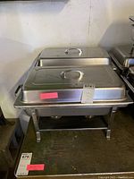 Photo of three chafing dishes stacked side by side showing their lids and overall structure with foldable legs and handles.