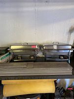 Front view of two stainless steel chafing dishes with covers on a black metal shelf. Several flat metal lids or trays stacked in front.