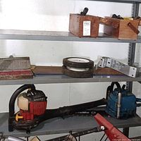 Shelf with the Homelite leaf blower (red and black), old blue SXL chainsaw, two rubber wheels, two wooden boxes, and two hand saws.