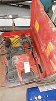 Open red Hilti case containing a Hilti TP800 chisel hammer, power cord, auxiliary handle, and yellow instruction manual.