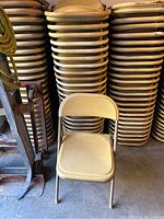 Stack of beige folding chairs with metal frames and padded seats and backs, showing signs of dirt, stacked against a wall.