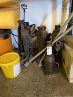 Four mechanical lifts and one hydraulic bottle jack with handles grouped together on concrete floor near wall.