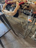 Three-quarter view of metal shelving unit showing top, middle, and bottom wire shelves, with some items on the top shelf (contents not included). Label with orange tag is visible on one leg.