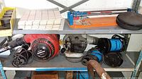 Shelving unit with sump pumps, leaf blowers, tiles, tile cutter, hockey pucks, and hand drill arranged on metal shelves.