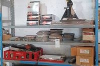 Full view of three metal shelves holding tire tubes in boxes, a mechanical jack stand, stacks of circular abrasive wheels, red crate with hand tools, and some cardboard boxes and sandpaper sheets.