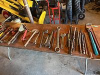 Photo of assorted large and medium wrenches, auger drill bits, and pipe wrenches on wooden table