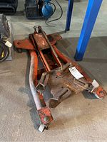 Side angle view of a heavy transmission jack showing caster wheels and lifting platform with adjustable brackets. Visible wear and rust on the orange painted metal.