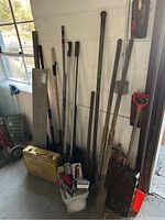 Photo of various yard tools including long-handled shovels, hole diggers, a large saw, and a yellow toolbox with hand tools and accessories in a bucket.