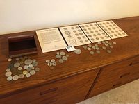 Setup showing rows of assorted coins laid out on a wooden surface including quarters in printed boards and other loose coins, plus a wooden box.