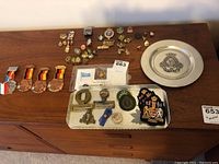 Overview of collection showing various military pins, badges, patches, belt buckles, and a pewter plate on a wooden surface.