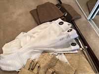 White sheer curtains and brown curtains with grommets laid on floor with curtain rods and gold looped top curtains partially visible