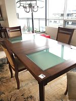 Dining table with black walnut frame, frosted glass inserts on the tabletop, and four matching wooden chairs with beige cushions around it.