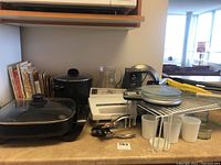 Multiple kitchen appliances and accessories including electric skillet, steamer, meal maker express, can opener, Magic Bullet blender, kitchen scale, and wine bottle opener arranged on a countertop.