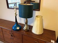 Photo showing both lamps on a wooden sideboard with one teal lamp shade attached and a cream fabric lamp shade separated.