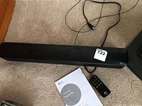 LG SK1 sound bar stretched out on carpet floor with attached power cord, remote control, and user manual beside it.