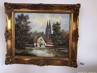 Front view of the oil painting in ornate gold frame showing European village scene with river and cathedral spires.