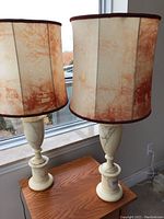Pair of alabaster table lamps on wooden surface near window showing entire lamps and shades.