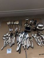 Overview photo showing multiple pieces of silver plated flatware (forks, spoons, knives) and some holloware items like candlesticks and covered dishes arranged on cardboard.