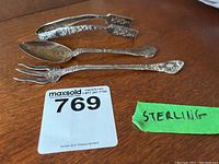 Three sterling silver serving utensils displayed together on wood surface, showing fork, spoon, and tongs with floral patterns and tarnish.