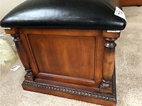 Side view of the ottoman showing black leatherette top and warm brown carved wooden base with turned column decoration