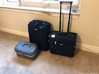 View of two black travel carry-on bags and a gray hard shell cosmetic case on carpet in front of window.