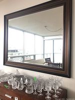 Photo of large wall mirror with dark brown wood frame reflecting a window and furniture in the room. Mirror mounted above a cabinet with crystal and glassware displayed.