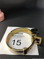 Gold-tone adjustable bangle bracelet shown from side angle on auction card with lot number 15 visible. The bracelet has a belt-like buckle closure and white enamel detailing on the exterior.