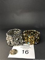 Two stretch bracelets, one silver with clear crystals and metal beads, one gold with amber rhinestones, placed on auction card with coin for size reference.
