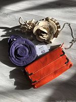 Three leather cross body bags arranged on a wooden floor with sunlight: tan round bag with leather flower, purple round bag with leather flower detail, and orange rectangular bag with black accents