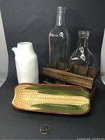 Hand-painted corn dish, white vase, and glass bottles in wooden holder against black background with coin for scale.