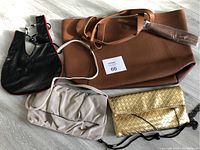 Four assorted handbags displayed on a floor including a large brown leather tote, black leather bag with red trim, light gray fabric bag, and gold woven clutch bag.
