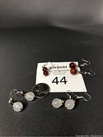 Four pairs of natural agate dangle earrings arranged around a MaxSold number card and a quarter coin for scale. Two pairs are red agate and two pairs are white agate, each with silver hooks.