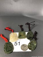 Photo of five jade pendants of varying green and yellow tones, each with a braided rope chain, displayed beside a quarter coin for scale and a lot tag '51'.